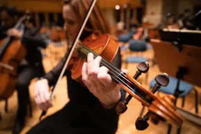 A imagem mostra uma musicista tocando uma viola muito de perto. Os dedos estão dedilhando o braço do instrumento, e o arco está tocando as cordas.