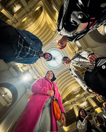 A imagem mostra quatro pessoal, duas mulheres e dois homens, olhando para a camera que está posicionada no chão. Ao fundo, é possivel ver o teto da Sala São Paulo e a cúpula colorida. 